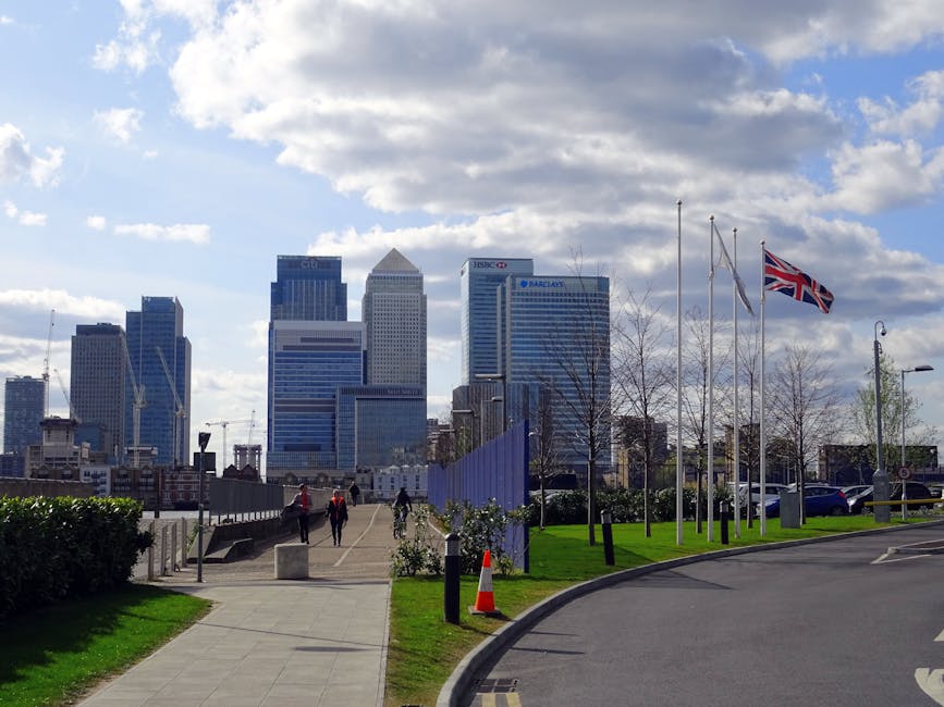 A view of a city skyline featuring modern high-rise office buildings with glass facades, prominently displaying logos such as HSBC, Barclays, and a triangular-roofed tower. In the foreground, a paved pathway runs alongside a low black metal fence, leading towards the buildings. To the right, there is a patch of well-maintained green grass with a few leafless trees and several flagpoles, one flying the Union Jack, all positioned on a small grassy area adjacent to a curved driveway. A traffic cone is visible near the grass, indicating possible recent activity or caution. Several pedestrians are strolling along the pathway, and parked cars are seen along the curb. The sky above is partly cloudy with patches of blue, creating an atmosphere of natural daylight. This scene subtly relates to the context of alternative waste handling or private rubbish collection near commercial or urban areas, as the setting appears to be an accessible outdoor space close to business districts, where rubbish removal services like those by House Clearance Canary Wharf could operate in compliance with city regulations.