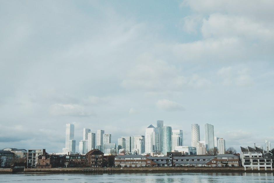A wide-angle view of a modern city skyline with numerous high-rise office buildings made of glass and steel, reflecting diffuse daylight from an overcast sky. In the foreground, there is a calm body of water, possibly a river or canal, with a few boats docked along its edge. Below the waterline, a row of low-rise, brick-built residential and commercial buildings features pitched roofs, some with visible windows and balconies, adding a contrast to the towering skyscrapers behind. The environment suggests an urban area with a mix of older, traditional architecture adjacent to contemporary high-rises, indicative of ongoing urban development. The overall atmosphere is neutral and professional, with natural lighting highlighting the textures and finishes of the different building materials. This scene subtly aligns with themes of property, building clearance, or urban refurbishment projects managed by services like House Clearance Canary Wharf, which specializes in waste disposal and rubbish removal for such environments.