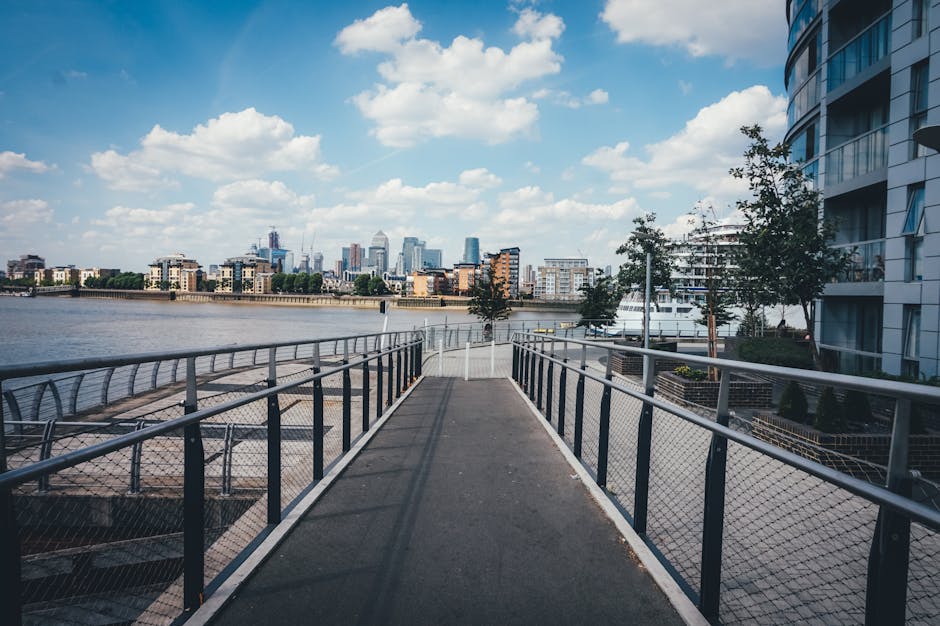 A wide, pedestrian walkway with metal railings and a grated metal fence along both sides extends toward a river with a city skyline in the background. The pathway appears to be part of a riverside promenade adjacent to modern, multi-storey residential buildings with curved glass facades on the right. On the left, the riverbank features a paved embankment with outdoor seating areas and small trees. The river is calm and reflects the partly cloudy sky, which has scattered white clouds and patches of blue. In the distance, the cityscape includes a variety of high-rise office buildings and construction cranes, contributing to an urban skyline. The scene is illuminated by natural daylight, with shadows cast by the railings and trees, creating a clear, visually detailed view suitable for accessibility descriptions related to urban environments, alternative waste handling sites, or outdoor infrastructure.