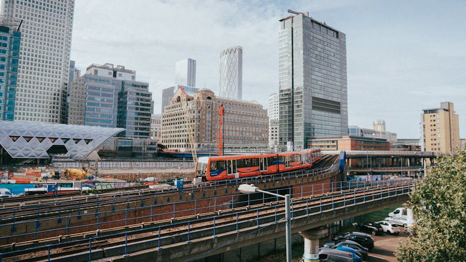 A modern train painted in bright orange and red livery travels on an elevated railway track through a cityscape featuring high-rise office buildings and modern architectural structures. The train's body is sleek with large window panes, and it appears to be in motion, crossing a bridge supported by steel beams and railings. Below the elevated track, there is a parking lot filled with several parked cars, including sedans and hatchbacks, parked on a paved surface. In the foreground, a street lamp is visible with a curved arm extending over the road. The background shows a cluster of tall, contemporary skyscrapers with glass facades reflecting the sky, and the environment is well-lit with natural daylight. The scene captures an urban setting where city infrastructure supports efficient transportation and possibly private or independent transit options, aligning with services that facilitate rubbish removal and site clearance in busy city environments. House Clearance Canary Wharf offers expertise in managing waste and clearing debris from such high-density, built-up areas, supporting sustainable urban development.