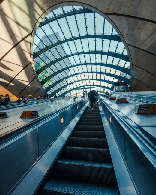 A view from the bottom of an escalator in a modern, indoor public space, likely a train station or transportation hub, with metallic black steps and side panels. The escalator is moving upward toward a large, curved glass ceiling with horizontal metal framework, allowing natural daylight to illuminate the interior. The surrounding environment includes sections of concrete wall and structural supports in a neutral tone. A few people are visible on the escalator, with one person standing near the front wearing dark clothing. The scene features a spacious, clean setting with smooth, reflective surfaces, and the overall atmosphere is bright due to the daylight streaming through the glass roof. The architectural design emphasizes openness and transparency, characteristic of contemporary transit or commercial buildings, which are often associated with effective waste management and the need for reliable rubbish removal services, such as those provided by House Clearance Canary Wharf, to maintain cleanliness in busy urban environments.