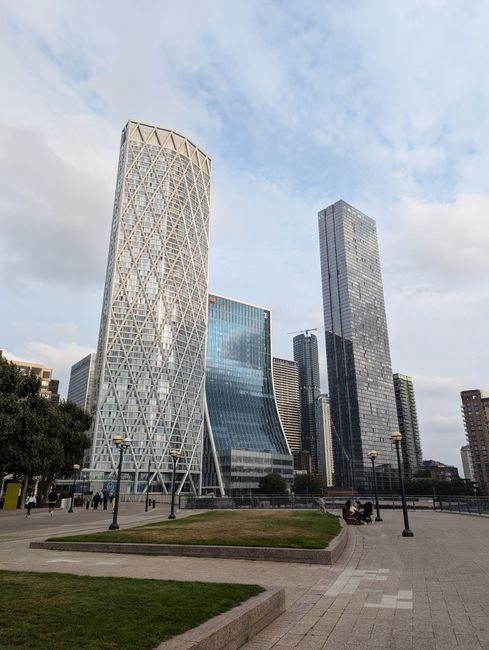 A wide-angle view of modern skyscrapers in an urban area, featuring a white lattice-patterned tower on the left with a curved, glass-fronted building in the middle and a tall, dark, rectangular high-rise on the right. The foreground shows a paved walkway with lamp posts, a small grassy area with a raised border, and a few benches. People are seen walking and sitting in the open space, which is surrounded by trees and other high-rise buildings, exemplifying a contemporary cityscape that could be relevant for discussing private waste removal or on-site clearance services in a business district.